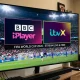 A smart TV screen in a living room displaying a split graphic of the BBC iPlayer and ITVX logos over a live World Cup 2026 football match.